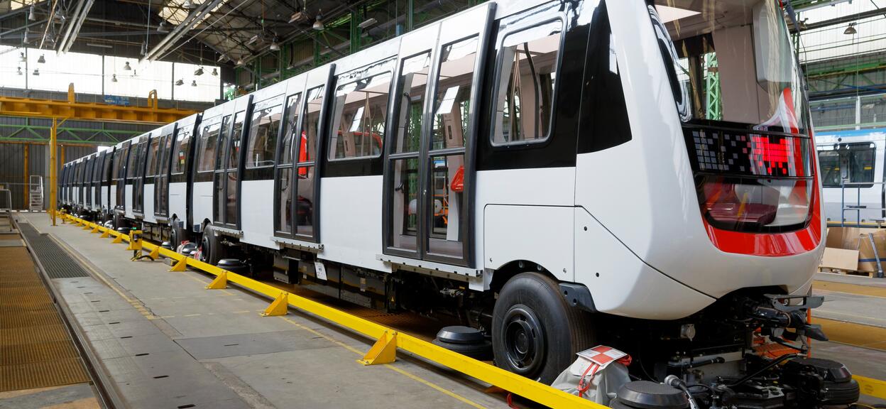 We see a metro train in an assembly hall. The metro is oriented from left to right (with the front on the right-hand side of the picture). The metro has a white livery (no logo), with the front end circled in orange. It is protected by a yellow rail that runs the length of the assembly line.
