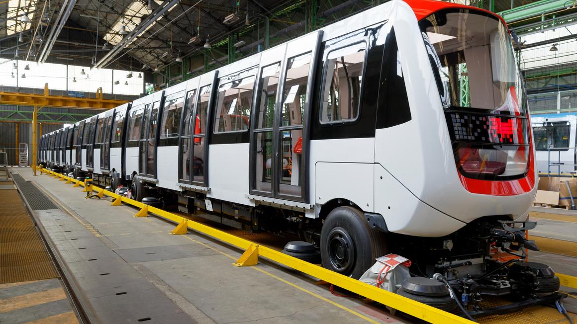 A 52-metre Lille metro trainset being assembled at Alstom’s Valenciennes-Petite Forêt site (© Alstom/Samuel Dhote) We see a metro train in an assembly hall. The metro is oriented from left to right (with the front on the right-hand side of the picture). The metro has a white livery (no logo), with the front end circled in orange. It is protected by a yellow rail that runs the length of the assembly line.