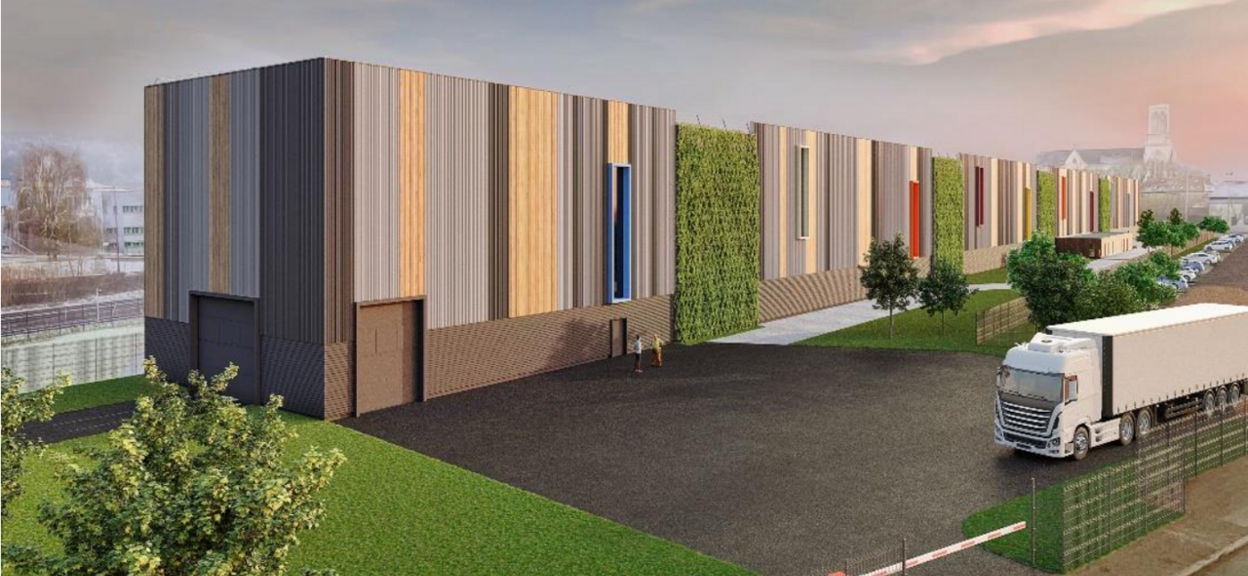 Un grand bâtiment traverse l’image de gauche à droite. Devant le bâtiment, deux personnes discutent sur un parking. Un camion blanc est garé en bas à droite de l’image. On voit des voitures stationnées plus loin et, dans le lointain, une église avec son clocher.