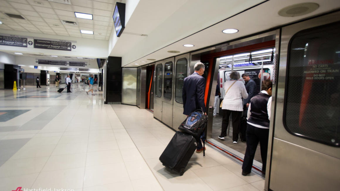 You can see the boarding platform of an airport shuttle. The floor is tiled with large light-coloured tiles. A passenger in a suit is preparing to cross the threshold of the shuttle, on the right, dragging behind him a wheeled suitcase and a backpack. Other passengers have already boarded the shuttle, whose doors are open, as are the station’s platform doors. Other passengers can be seen waiting or moving in the background. Overhead signage can be seen at the top as well as a video screen above the entrance