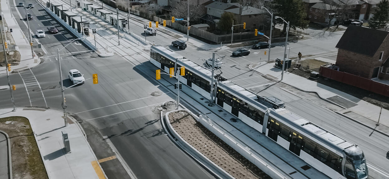 The image shows a modern urban intersection featuring a dedicated tramway line. At the centre, a sleek articulated tram in white and black runs on segregated tracks bordered by concrete curbs. The tram, composed of multiple cars, is equipped with pantographs for overhead electric power supply. On the left, there is a station with raised platforms, sheltered by metallic structures and fitted with information panels. Several traffic lights and road signs regulate the movement of vehicles and pedestrians.