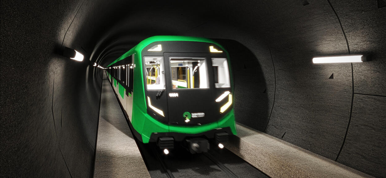 The image shows a modern metro train travelling through a dark tunnel, lit by longitudinal lights fixed to the walls. The train, with its resolutely contemporary design, has a green and black livery. The sleek, angular front incorporates white LED lighting in the form of sharp lines, with the words “Suburban Rail Loop” in white lettering. Through the large windows in the cab and cars, you can see the train’s bright interior.