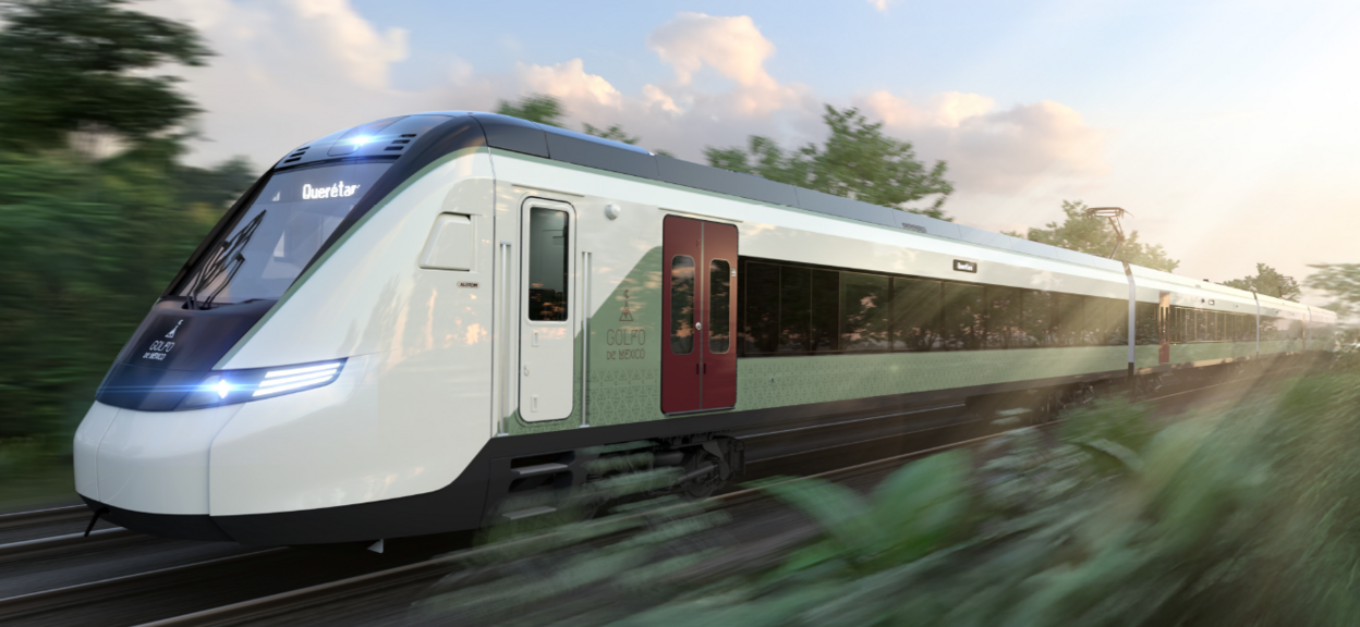 L’image montre un train moderne filant à vive allure sur une voie ferrée entourée de végétation luxuriante. Au premier plan, le train occupe la majorité de l’image : sa forme aérodynamique et ses lignes épurées évoquent une conception contemporaine. La motrice présente une face avant arrondie, soulignée par des éclairages LED bleu clair qui renforcent son aspect technologique. Sur le côté, la livrée combine des tons blancs, gris et verts.