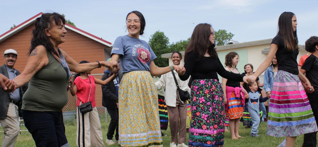 The image shows a group of people gathered outside on a lawn, participating in what appears to be a group dance or community activity. The participants are arranged in a circle or line, holding hands. Several women are wearing long, colourful skirts with floral or geometric patterns and bright colours (yellow, blue, pink, green) reminiscent of traditional dress. Adults, as well as a few children, are participating or observing the scene. Some people are wearing more modern outfits.
