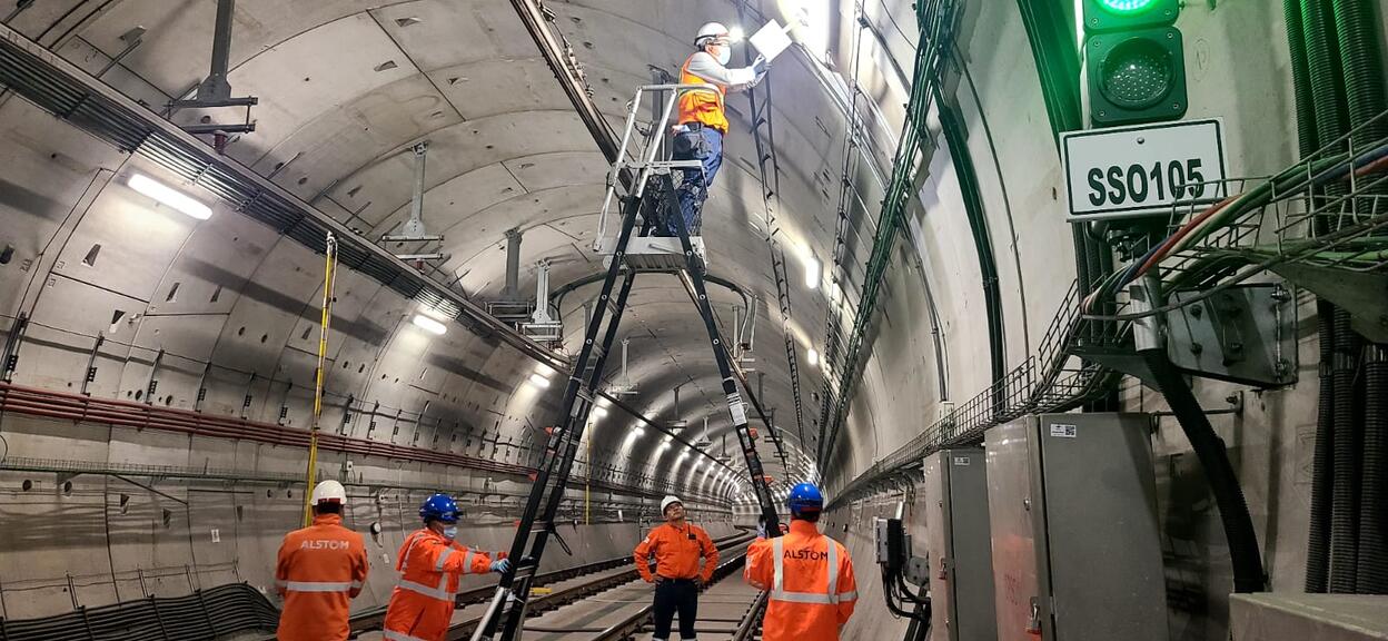 L’image montre une équipe de techniciens travaillant à l’intérieur d’un large tunnel ferroviaire. Les parois en béton, parfaitement courbées, s’étendent en perspective et sont éclairées par une série de luminaires fixés au plafond. Au centre, un technicien en gilet orange et casque de sécurité se tient en hauteur sur une grande échelle articulée afin d’intervenir sur un boîtier ou un élément électrique situé en hauteur.
