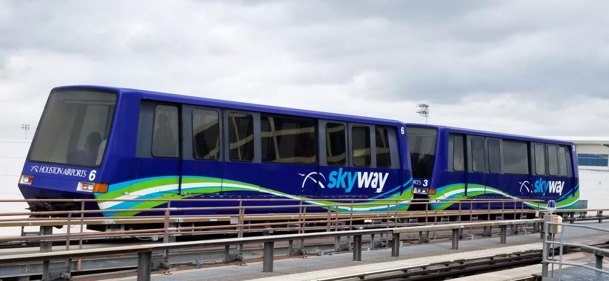 The image shows an automated people-mover travelling along an elevated track. The train consists of two dark blue cars, decorated with wavy green and turquoise lines. On the sides, the word ‘Skyway’ appears in white letters, accompanied by a pictogram depicting an aerial trajectory. At the front, a ‘Houston Airports’ sign indicates that this is an internal shuttle system used to connect different terminals at Houston Airport. The cars have large tinted windows.