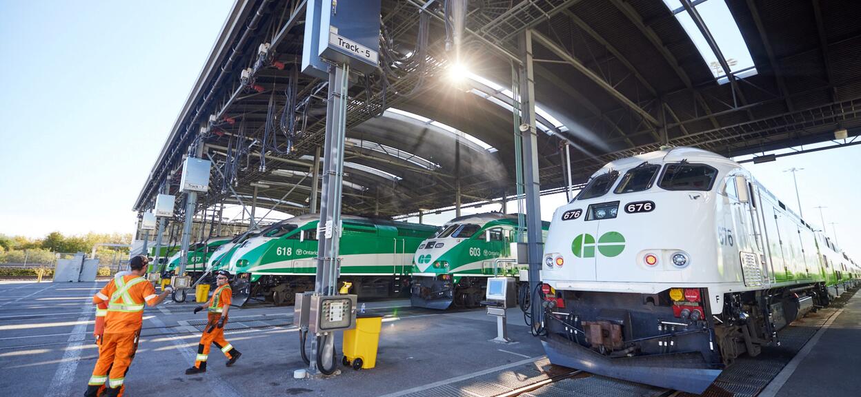 The image shows a vast railway depot where several locomotives are lined up beneath a large metal structure. In the foreground, a white and green locomotive is parked on a track, with its number visible at the front (676). Other locomotives, painted mainly green with white stripes, are on the adjacent tracks; they all bear the ‘GO’ logo at the front. The roof of the depot, open at the sides, lets in sunlight, which filters through the structure and creates bright reflections on the trains.