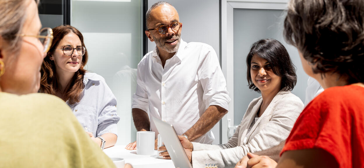 The image shows a business meeting taking place in a bright, contemporary setting. Several people are gathered around a high table with a white top. They are placed in a workspace with light-coloured, glass-panelled walls.  In the centre of the photo, a person standing, dressed in a white shirt and wearing glasses, appears to be speaking or presenting something. This person is leaning slightly towards the table and looking at one of the participants, giving the impression of a direct exchange.
