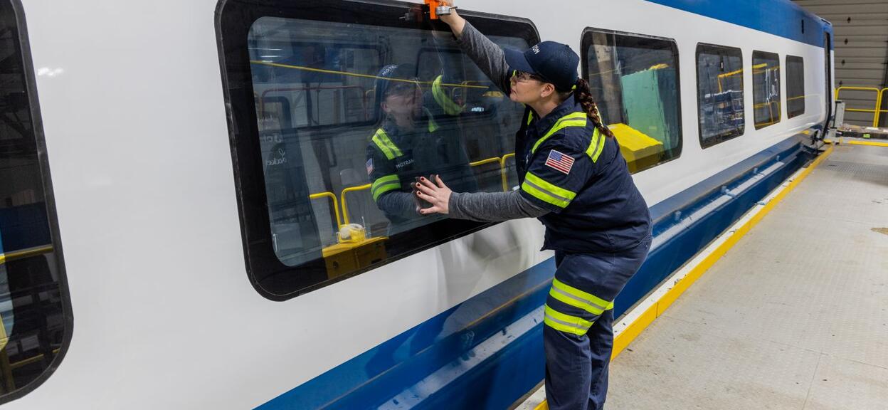 The image shows a scene taking place inside a railway workshop. In the foreground, a person in work clothes is wearing a dark uniform with fluorescent yellow reflective strips and a badge featuring an American flag on the sleeve. This person is standing next to a white and blue train and is using an orange tool to work on a large rectangular window. One hand is holding the surface of the window whilst the other is handling the tool.