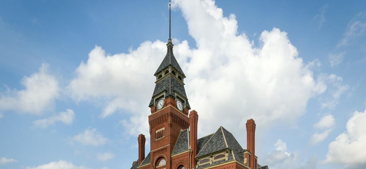 The image shows a historic-style red-brick building, photographed in broad daylight under a blue sky dotted with white clouds. The main structure is dominated by a central tower, topped by a pyramidal bell tower and a slender metal spire. Halfway up the tower, two circular clocks are visible on opposite sides. The facade features decorative architectural elements, including arches above the windows, light-coloured stone frames, and several vertical brick chimneys.