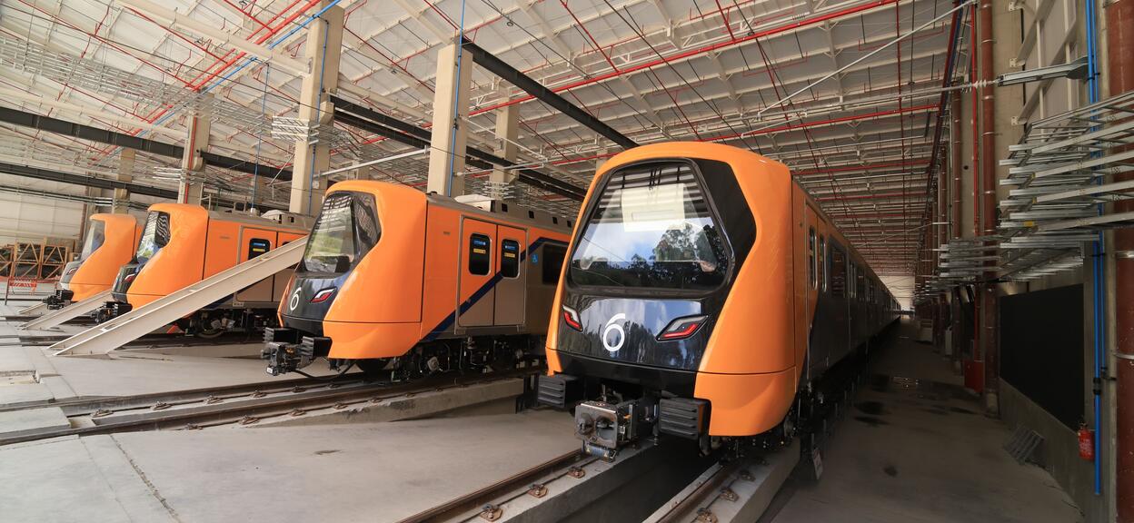 This picture shows several metro trains parked inside a large maintenance centre or railway depot. In the foreground, an orange and black train is positioned on a sidings track. Its design is modern, featuring a sleek black front end (in which the natural surroundings outside the site are reflected), a wide, dark windscreen and lights integrated into the dsign. A logo symbolising the number 6 stands out between the lights.