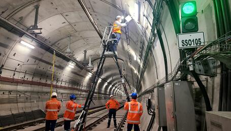 L’image montre une équipe de techniciens travaillant à l’intérieur d’un large tunnel ferroviaire. Les parois en béton, parfaitement courbées, s’étendent en perspective et sont éclairées par une série de luminaires fixés au plafond. Au centre, un technicien en gilet orange et casque de sécurité se tient en hauteur sur une grande échelle articulée afin d’intervenir sur un boîtier ou un élément électrique situé en hauteur.