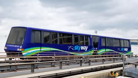 The image shows an automated people-mover travelling along an elevated track. The train consists of two dark blue cars, decorated with wavy green and turquoise lines. On the sides, the word ‘Skyway’ appears in white letters, accompanied by a pictogram depicting an aerial trajectory. At the front, a ‘Houston Airports’ sign indicates that this is an internal shuttle system used to connect different terminals at Houston Airport. The cars have large tinted windows.