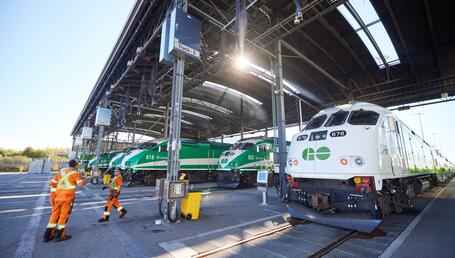 The image shows a vast railway depot where several locomotives are lined up beneath a large metal structure. In the foreground, a white and green locomotive is parked on a track, with its number visible at the front (676). Other locomotives, painted mainly green with white stripes, are on the adjacent tracks; they all bear the ‘GO’ logo at the front. The roof of the depot, open at the sides, lets in sunlight, which filters through the structure and creates bright reflections on the trains.