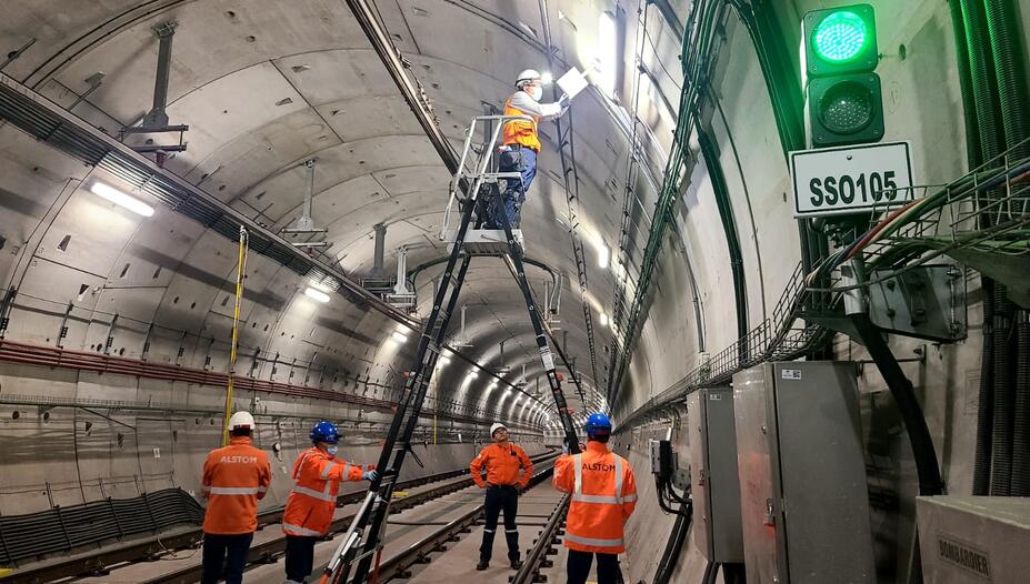 L’image montre une équipe de techniciens travaillant à l’intérieur d’un large tunnel ferroviaire. Les parois en béton, parfaitement courbées, s’étendent en perspective et sont éclairées par une série de luminaires fixés au plafond. Au centre, un technicien en gilet orange et casque de sécurité se tient en hauteur sur une grande échelle articulée afin d’intervenir sur un boîtier ou un élément électrique situé en hauteur.