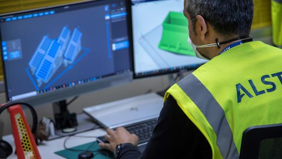 Employee in yellow Alstom logo vest typing at computer with 3D models on screen