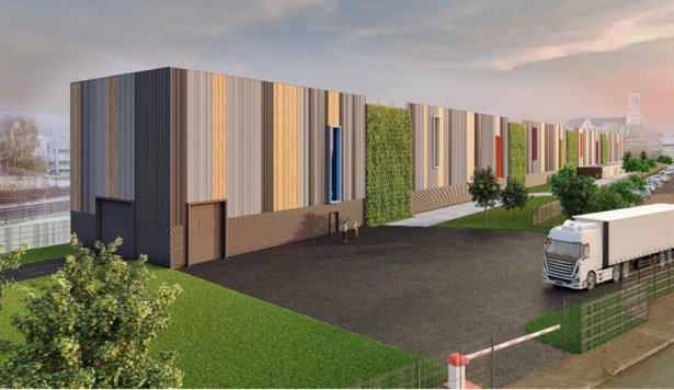 Un grand bâtiment traverse l’image de gauche à droite. Devant le bâtiment, deux personnes discutent sur un parking. Un camion blanc est garé en bas à droite de l’image. On voit des voitures stationnées plus loin et, dans le lointain, une église avec son clocher.