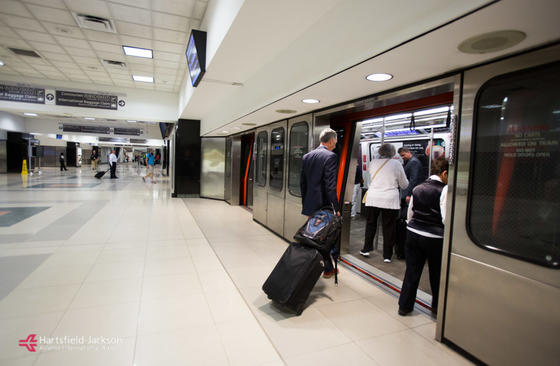 You can see the boarding platform of an airport shuttle. The floor is tiled with large light-coloured tiles. A passenger in a suit is preparing to cross the threshold of the shuttle, on the right, dragging behind him a wheeled suitcase and a backpack. Other passengers have already boarded the shuttle, whose doors are open, as are the station’s platform doors. Other passengers can be seen waiting or moving in the background. Overhead signage can be seen at the top as well as a video screen above the entrance